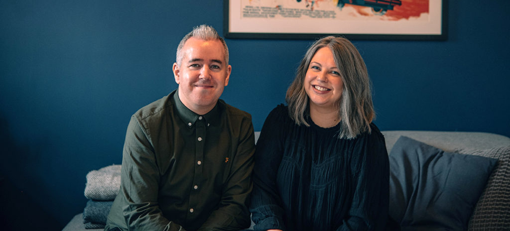 A photo of Jonny and Bex Rae-Evans sat together, smiling nervously because they hate having their photo taken. They're sat on the sofa, and behind them is a deep blue wall, with a Back to the Future 3 print hanging on it.